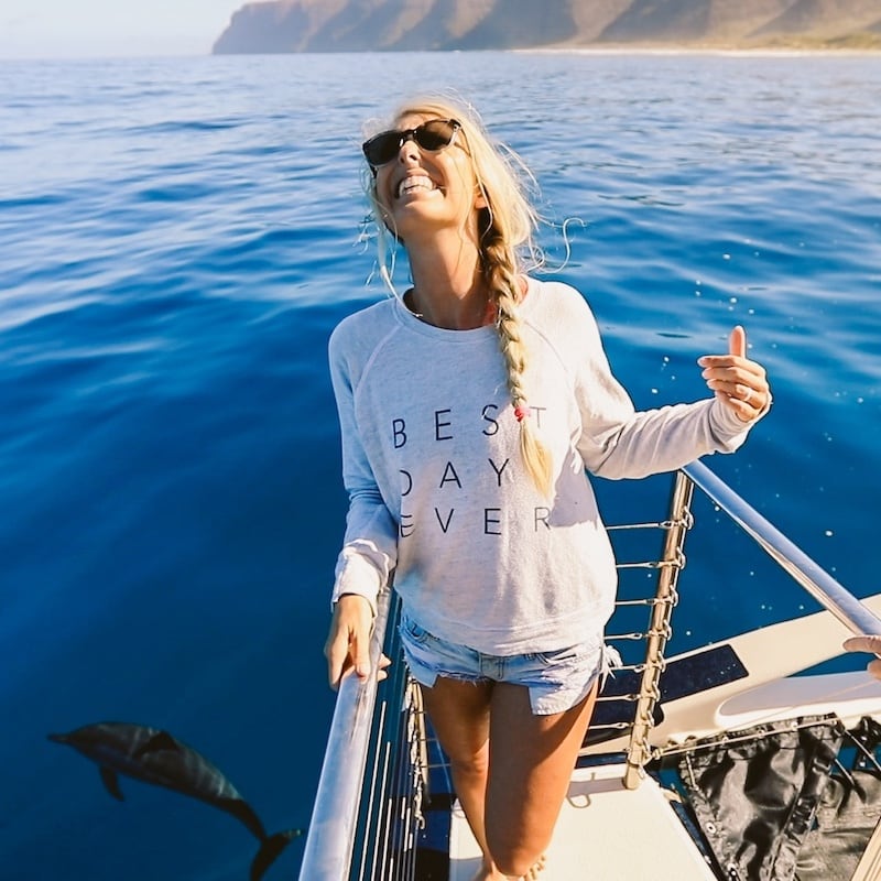 Woman smiling and making a shaka sign on the bow of a Kauai catamaran with a dolphin swimming in the background.