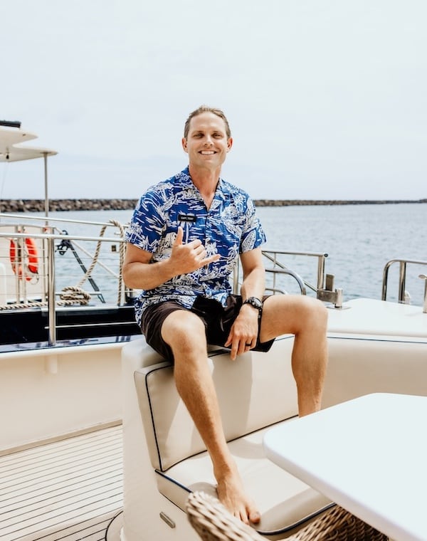 A crew member of Captain Andy's in a blue Hawaiian shirt making a shaka sign while sitting on a boat docked at the harbor, with the ocean in the background.