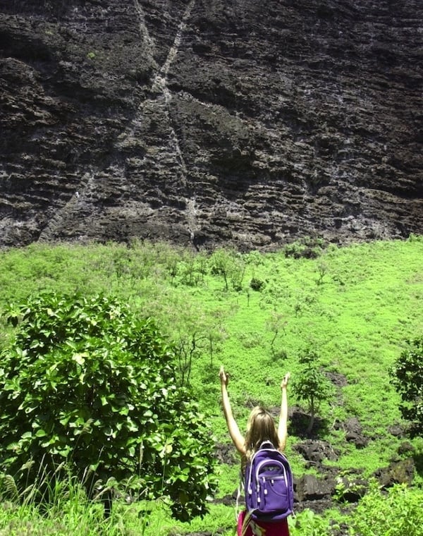 Hiker with a purple backpack raising arms in triumph in front of a towering, lush green cliff.